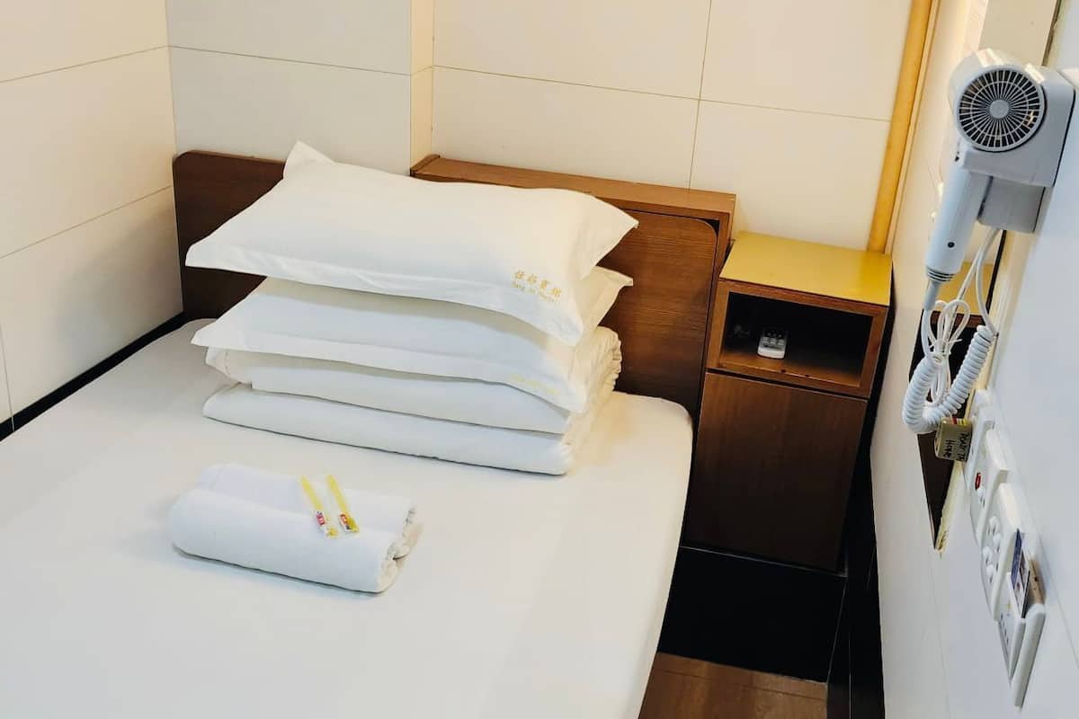 A neatly made bed with white linens, a wooden headboard, a bedside table with a remote control, and a hairdryer on the wall.