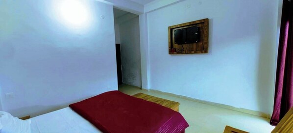 A room with a bed, a wooden TV mount on the wall, and a doorway leading to another room.