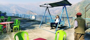 A terrace with a swing, green chairs, and tables, overlooking a mountainous landscape. - The Yamuna Deck Resort and Cafe (Dhanaulti)
