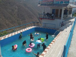 Una piscina con gente y anillos inflables, rodeada por un suelo a cuadros y vista a un paisaje montañoso.