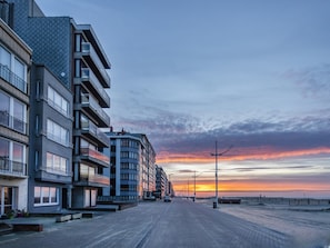 A modern multi-story building with balconies, a paved walkway, and a sunset view.
