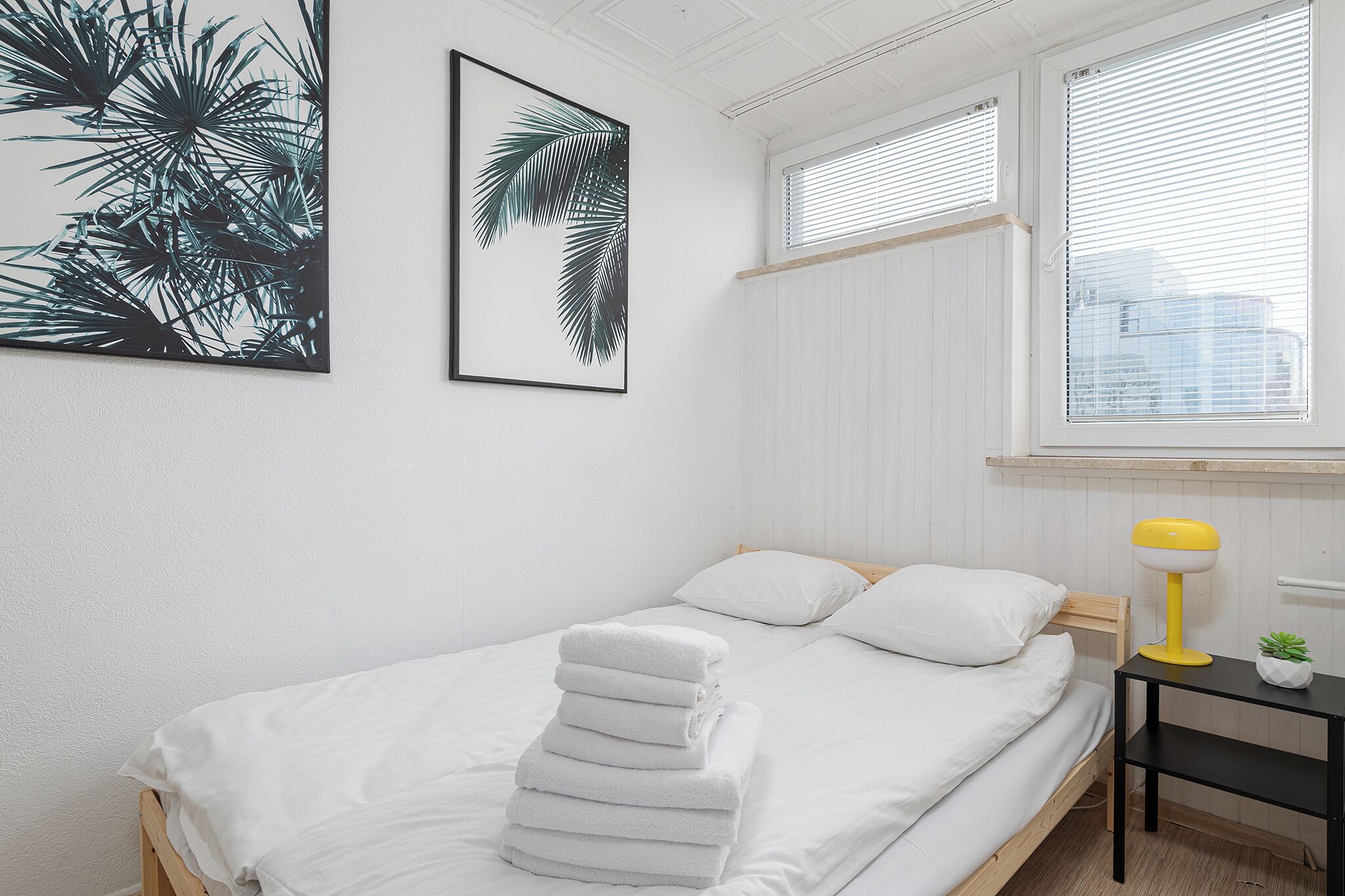 Quarto com cama, roupa de cama branca, pilha de toalhas brancas, abajur amarelo sobre uma mesa de cabeceira, pequena planta em vaso e grande quadro com folhas de palmeira na parede.