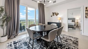 A modern hotel room with a round dining table, grey upholstered chairs, a potted plant, and a large window with curtains. - RIVER FLY-IN: Luxury Condo w/ Views & Amenities (Merritt Island)
