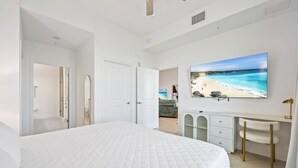 A bedroom with a large bed, a desk, a chair, a TV, and a view of a beach. - RIVER FLY-IN: Luxury Condo w/ Views & Amenities (Merritt Island)