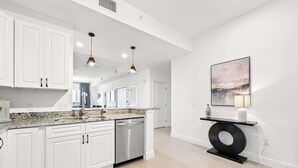 A modern kitchen with white cabinets, granite countertops, stainless steel appliances, pendant lighting, and a minimalist decor including a large abstract painting and a unique black console table. - RIVER FLY-IN: Luxury Condo w/ Views & Amenities (Merritt Island)