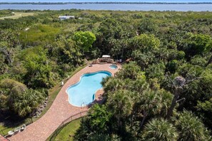 A large swimming pool surrounded by lush greenery, with a paved walkway leading to it. - RIVER FLY-IN: Luxury Condo w/ Views & Amenities (Merritt Island)