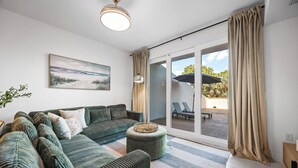 Un salon moderne avec un canapé vert, un pouf rond et une grande fenêtre donnant sur un espace extérieur aménagé.