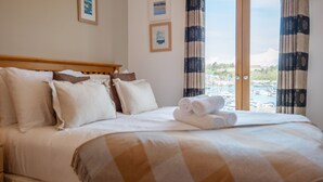 A neatly made bed with a checkered blanket, a wooden headboard, and a view of a marina through large windows. - Coastal Escape with Balcony & River Dart Views (Kingswear)