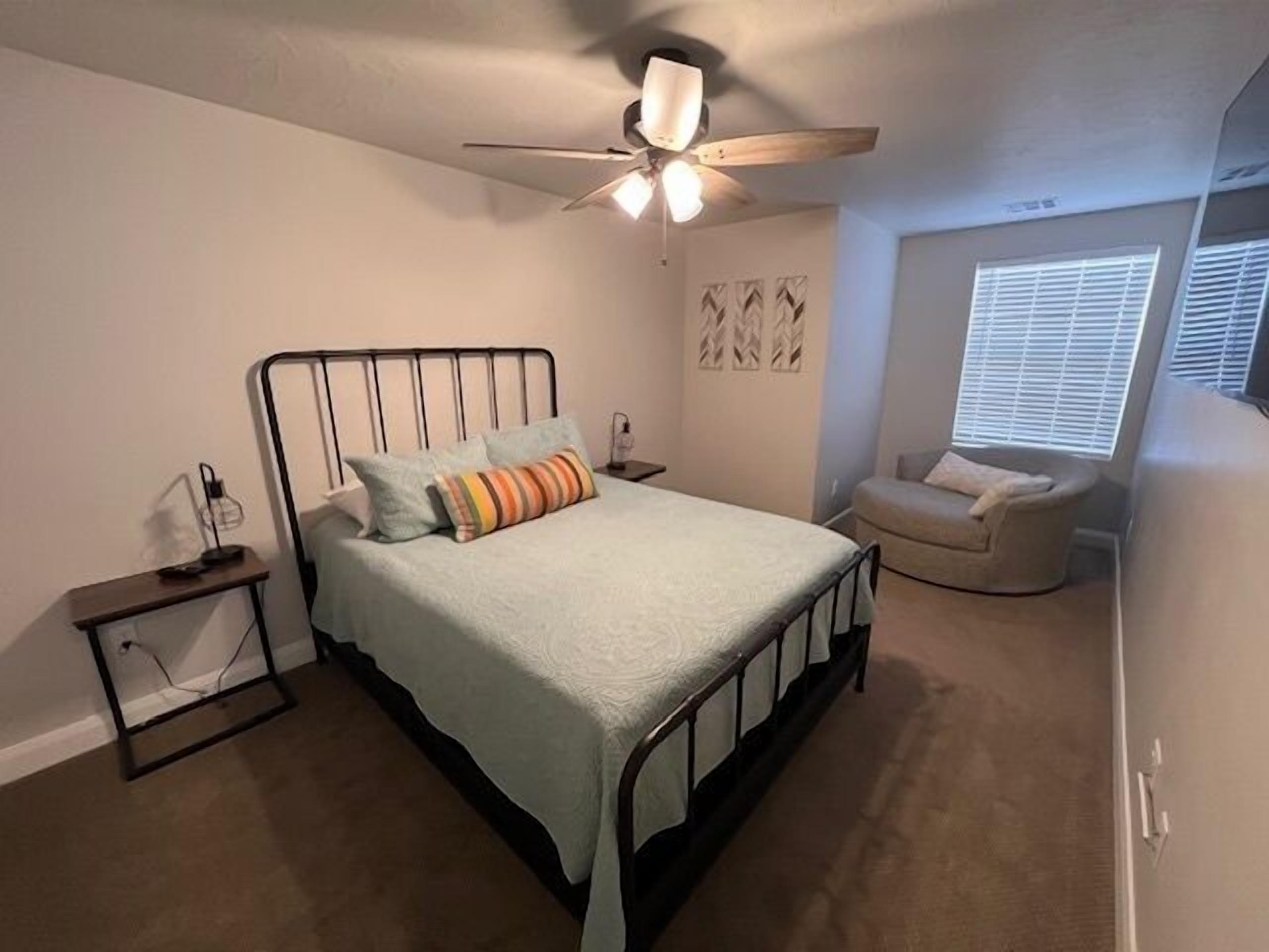 A bedroom with a bed, a nightstand, a chair, a ceiling fan, and a window with blinds.