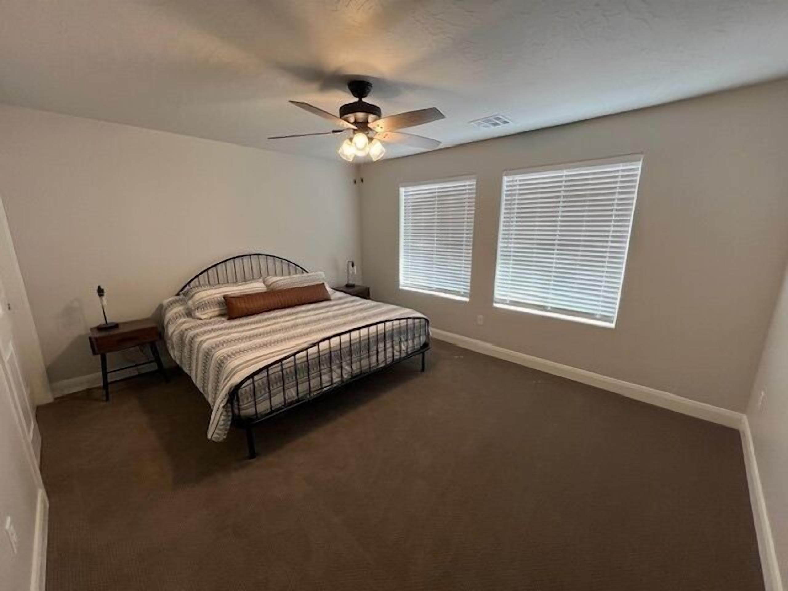 A bedroom with a bed, a ceiling fan, and two windows with blinds.