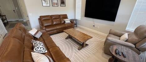Una sala de estar con un sofá de cuero marrón, una mesa de centro de madera, un sillón beige y un televisor montado en la pared.