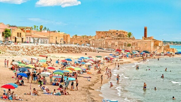 A crowded beach with people sunbathing and swimming, surrounded by historic buildings and a clear sky. - Nicuzza House (Avola)