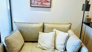 A beige sofa with striped and solid pillows, a framed picture of palm trees on the wall, and a floor lamp to the right.
