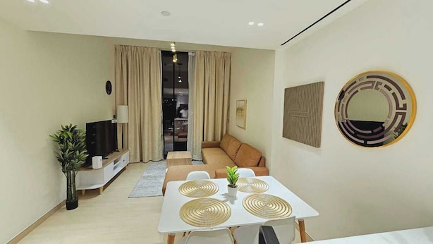 A modern living room with a sofa, a dining table with placemats, a TV on a stand, and a large mirror on the wall. - OYO HOME 1405 Spacious 1bed In Jvc Be (Dubai)