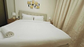A neatly made bed with white linens and a rolled towel, a headboard, two bedside lamps, and a wall-mounted light fixture. - OYO HOME 1405 Spacious 1bed In Jvc Be (Dubai)