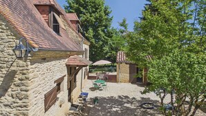 A stone building with a tiled roof, outdoor seating, and a tree in a courtyard.
