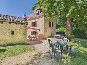 A stone building with a tiled roof, outdoor seating with a table and chairs, and a large tree in the background.
