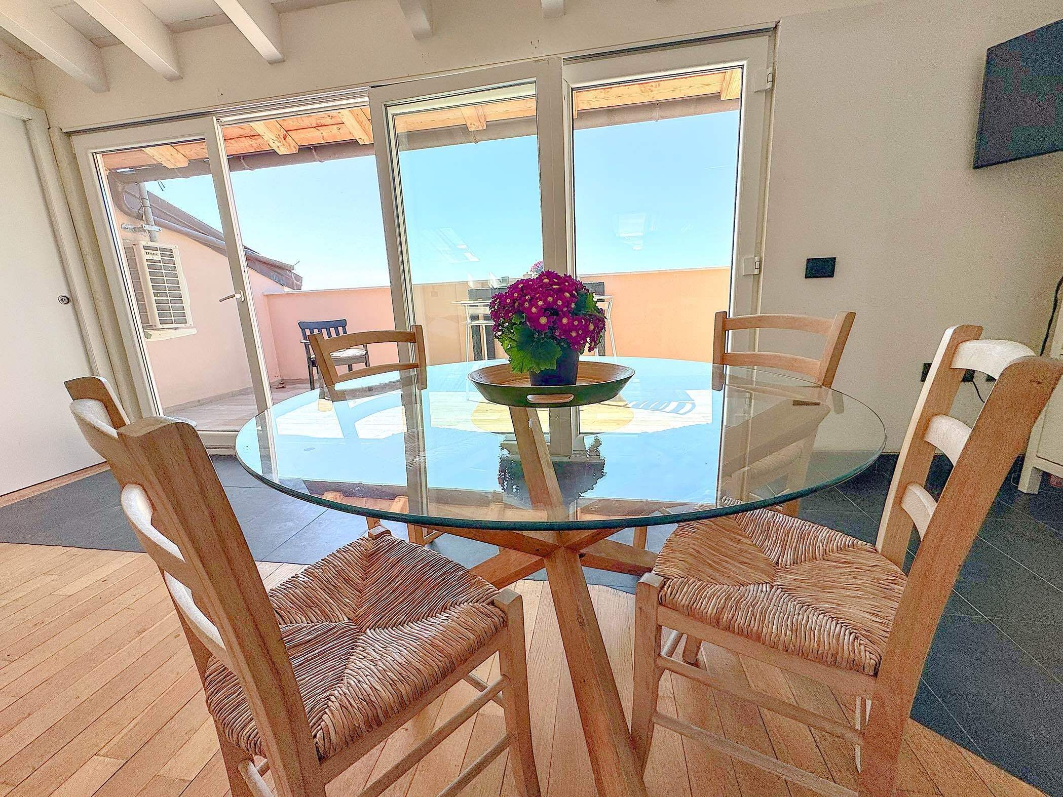 A round glass table with four wicker chairs, a potted plant, and a view of the outdoors through large sliding glass doors.