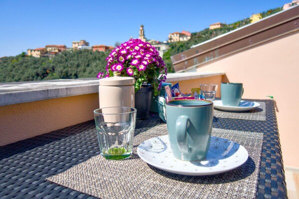 A table set with a cup, saucer, glass, and a potted plant on a balcony with a view of buildings and greenery.