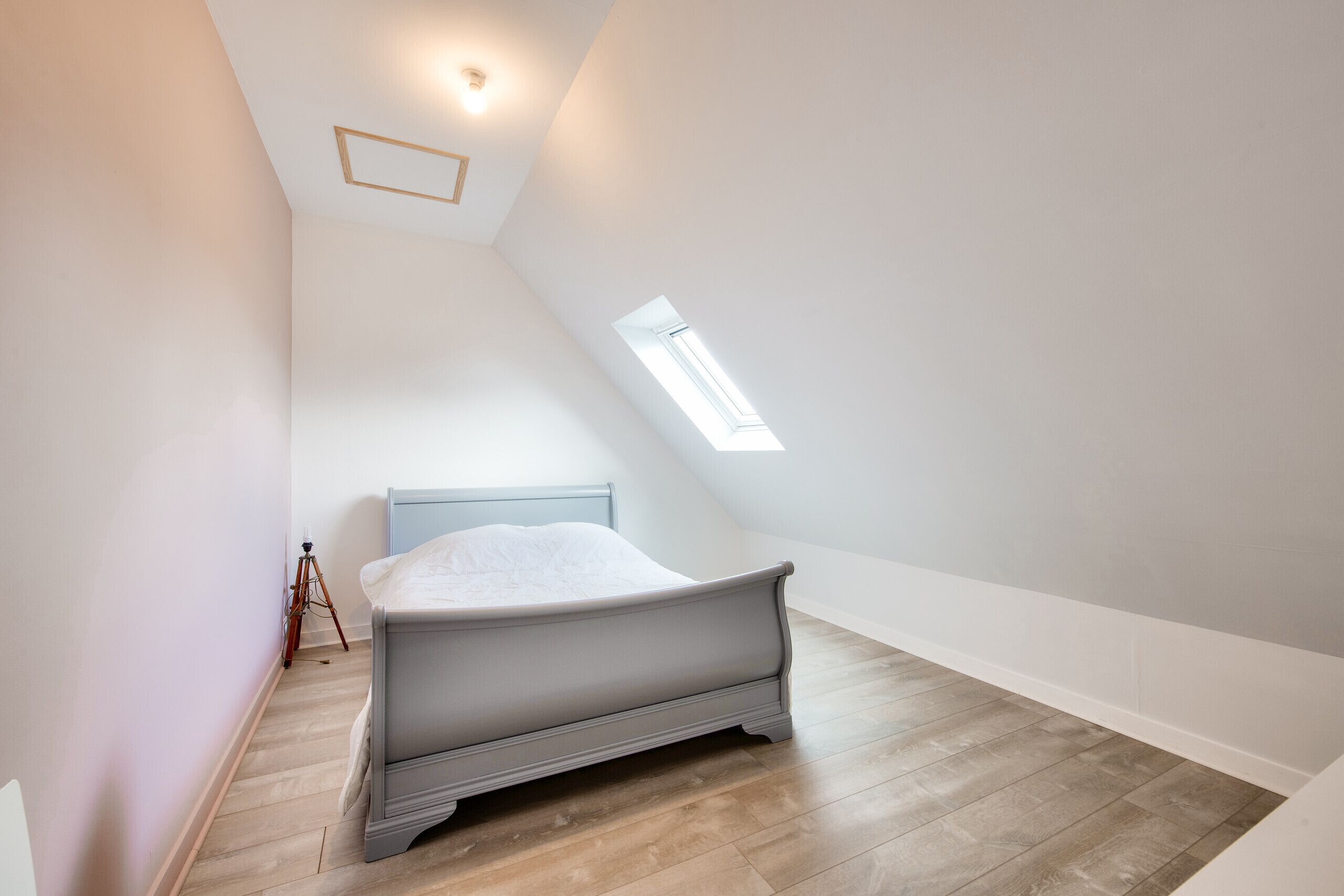 A bedroom with a sloped ceiling, a skylight, a bed, and a tripod.