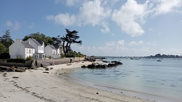 Un paysage côtier avec une plage de sable, un bâtiment blanc doté d’un balcon et une mer calme où voguent des bateaux.