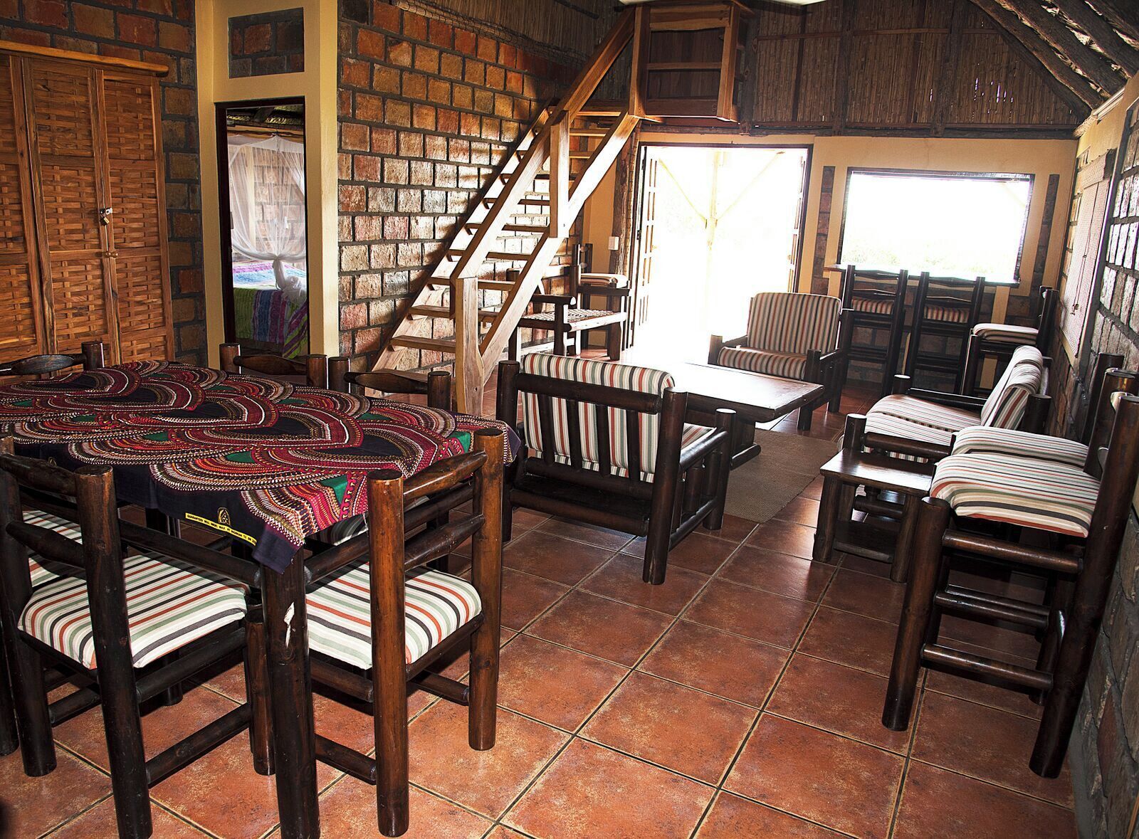 Un interior rústico con pared de ladrillo, escalera de madera y zona de comedor con sillas a rayas y mantel estampado.