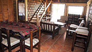 Un interior rústico con pared de ladrillo, escalera de madera y zona de comedor con sillas a rayas y mantel estampado.