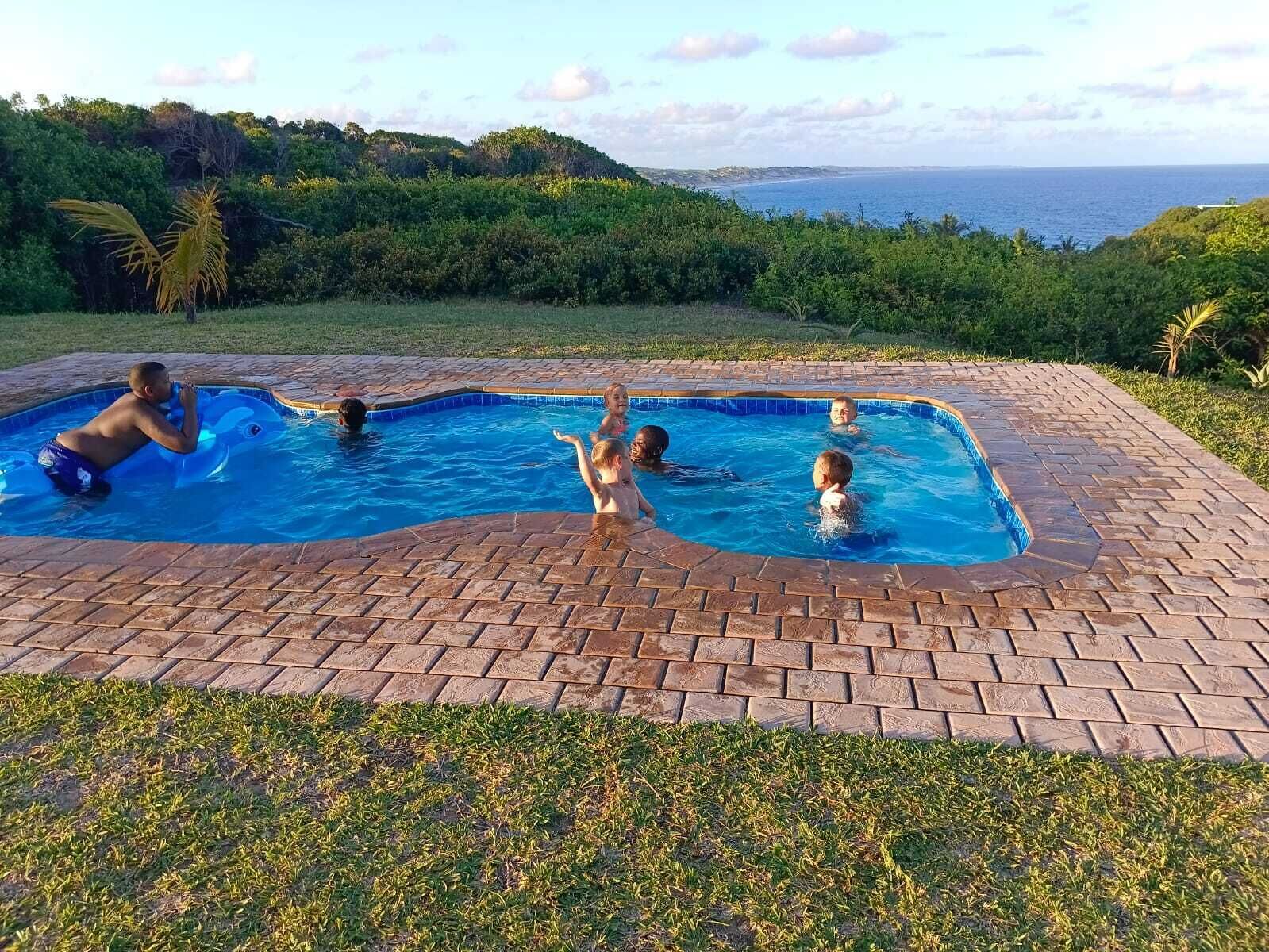 A swimming pool with a brick border, surrounded by grass and hills.