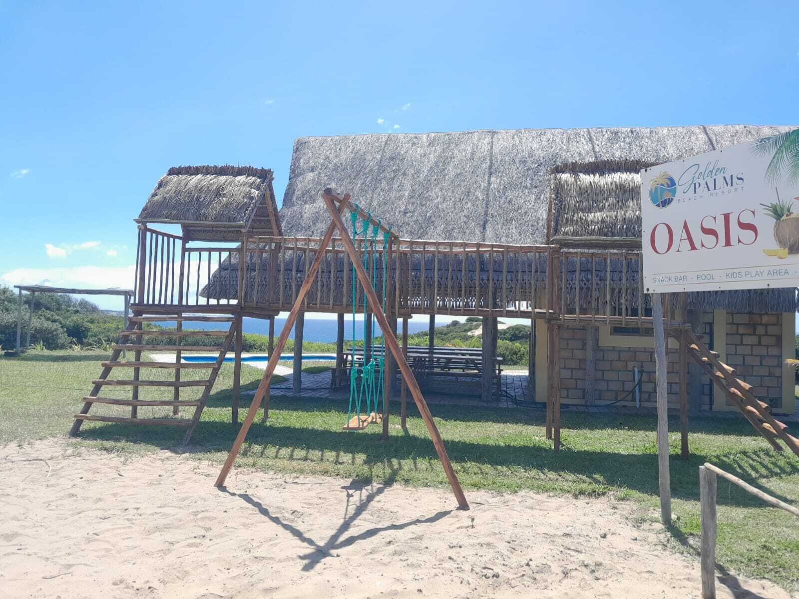 Een strandspeeltuin met een schommelset, een constructie met rieten dak en een bord voor Golden Palms Oasis met opsomming van voorzieningen.
