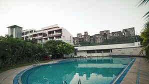 A swimming pool with a curved edge, surrounded by a concrete deck. In the background, there is a multi-story building with balconies and a garden area with trees. - BNGV Grandeur Hotel & Banquets (Bengaluru)