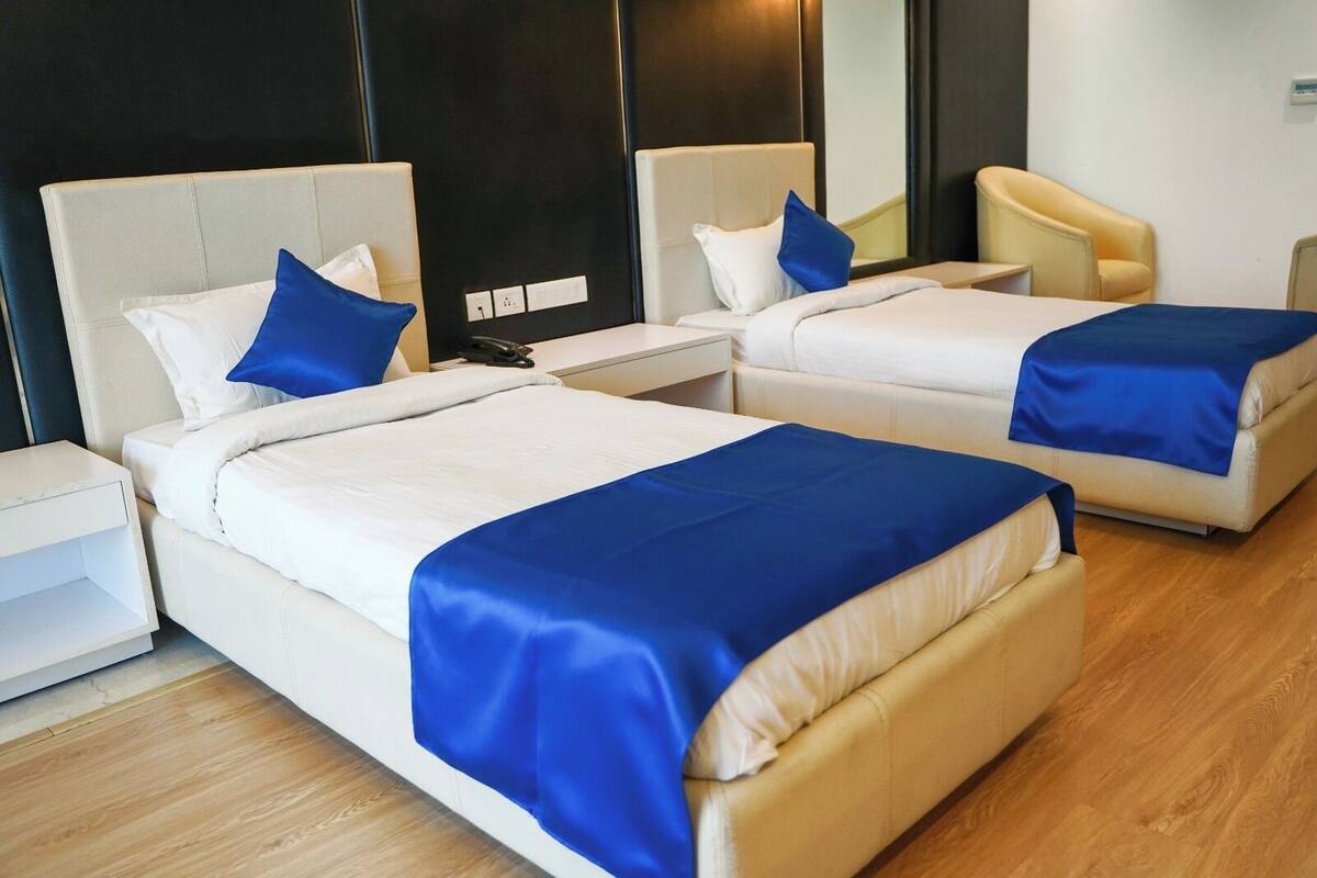 A hotel room with two beds, each with a blue bedspread, white pillows, and a bedside table.