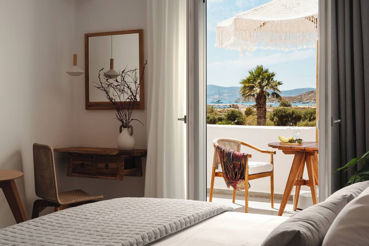 Une chambre avec un lit, un bureau et une chaise, un balcon avec une table et des chaises, et une vue sur la mer et les collines.