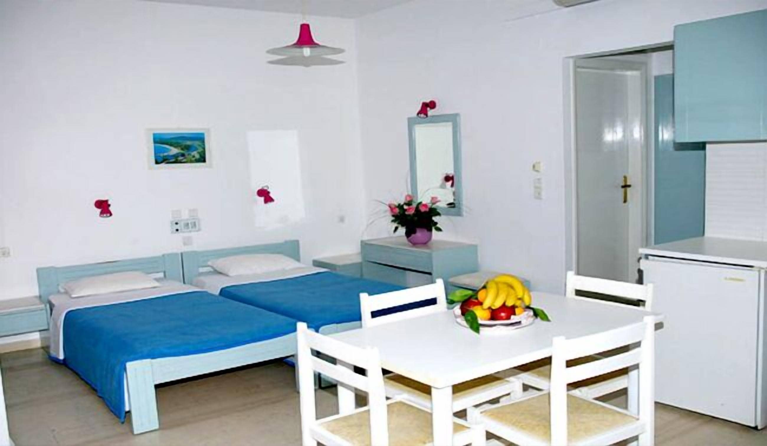Une petite chambre propre, avec deux lits, une table blanche et des chaises, un petit réfrigérateur et un miroir.