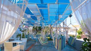 Een overdekt terras met hangstoelen en tafels, omringd door planten en een blauwe dakconstructie.