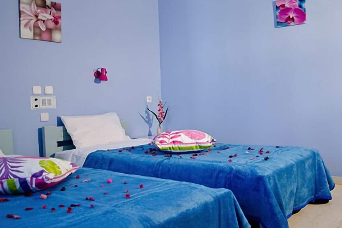 Une chambre avec deux lits, chacun recouvert d’une couverture bleue et agrémenté d’un coussin coloré. On y trouve des interrupteurs fixés au mur et un tableau encadré accroché au mur.