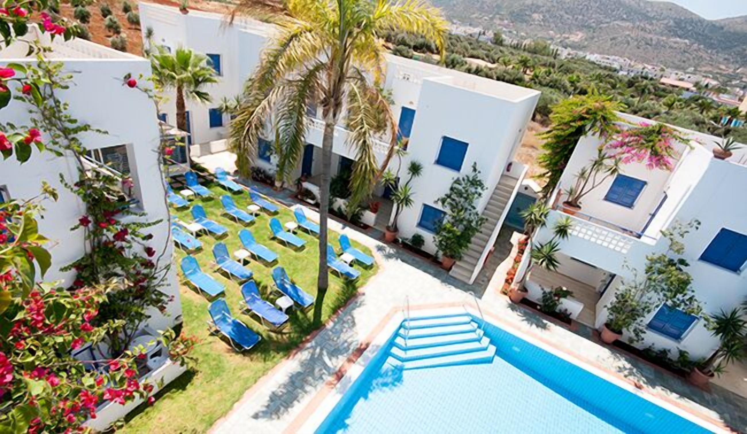Zona de piscina con sillas reclinables azules, una piscina y edificios blancos con ventanas azules.