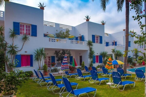 A courtyard with blue lounge chairs, white buildings with blue shutters, and potted plants. - Galini Apartments (Malia)