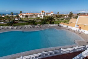 A large, clear swimming pool with lounge chairs and umbrellas, surrounded by a white fence. In the background, there is a large hotel with a red roof and palm trees.