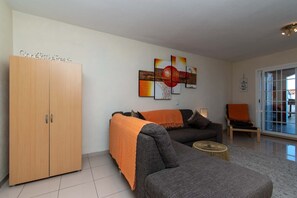 Sala de estar com sofá, mesa de centro e armário. Há um quadro na parede e uma janela com persianas.