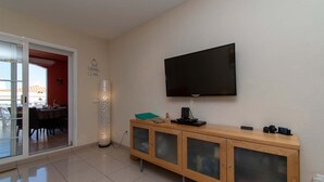 Una sala de estar moderna con un televisor de pantalla plana montado en la pared, un mueble de madera con puertas de vidrio y un área de comedor visible a través de una puerta de cristal.