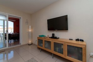 Een moderne woonkamer met een flatscreen televisie aan de muur, een houten kast met glazen deuren, en een eethoek zichtbaar door een glazen deur.