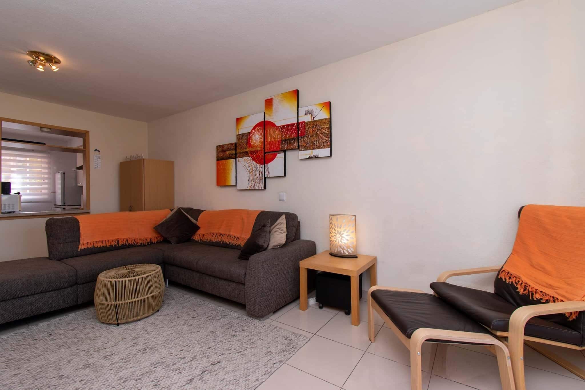 Ein modernes Wohnzimmer mit einer Eckcouch, einem kleinen runden Hocker, einem Beistelltisch mit Lampe und einem Sessel mit orangefarbener Decke.