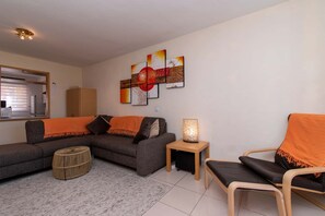Ein modernes Wohnzimmer mit einer Eckcouch, einem kleinen runden Hocker, einem Beistelltisch mit Lampe und einem Sessel mit orangefarbener Decke.
