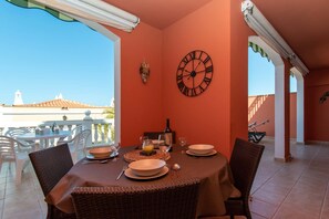 A dining area with a table set for two, a wall clock, and a view of a building with a tiled roof. - El Veril del Duque 13 - Two Bed (Costa Adeje)
