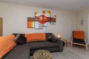 A living room with a grey sofa, orange throw, and a multi-panel artwork above it.