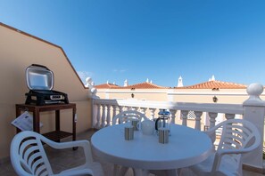 Een balkon met een ronde tafel en stoelen, een televisie en een klein karretje met een dienblad.