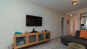 Una sala de estar con un televisor de pantalla plana montado en la pared, un mueble de madera con puertas de vidrio, un sofá gris y un otoman tejido.