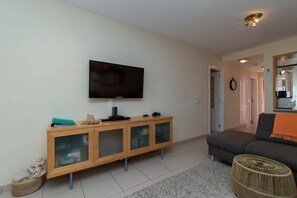 A living room with a flat-screen TV mounted on the wall, a wooden cabinet with glass doors, a grey sofa, and a woven ottoman. - El Veril del Duque 13 - Two Bed (Costa Adeje)