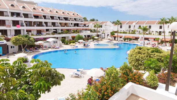 A hotel with a large swimming pool surrounded by lounge chairs and umbrellas, with multiple balconies and a tropical garden.