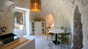Une pièce rustique aux murs de pierre, avec une table et des chaises, un luminaire suspendu et une vue sur un espace extérieur, accessible par une porte ouverte.